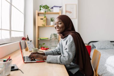 Başörtüsü takmış, evde dizüstü bilgisayar kullanarak gülümseyen siyah bir kız öğrenci. Genç Afrikalı bir kadın odasında öğreniyor ya da çalışıyor. Eğitim ve teknoloji kavramı.