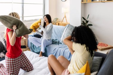 Pijama giyen neşeli çok ırklı bayan arkadaşlar pijama partisinde eğleniyorlar, evde yastıklarla oynuyorlar. Kadın arkadaşlığı ve gençlik kavramı