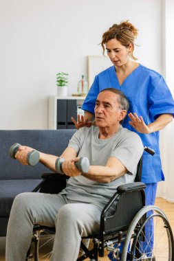 Tekerlekli sandalyede fizyoterapistle evde dambıl kullanırken çekilmiş dikey bir fotoğraf. Kadın terapist, huzurevi hizmetlerinde kıdemli adama yardımcı oluyor..