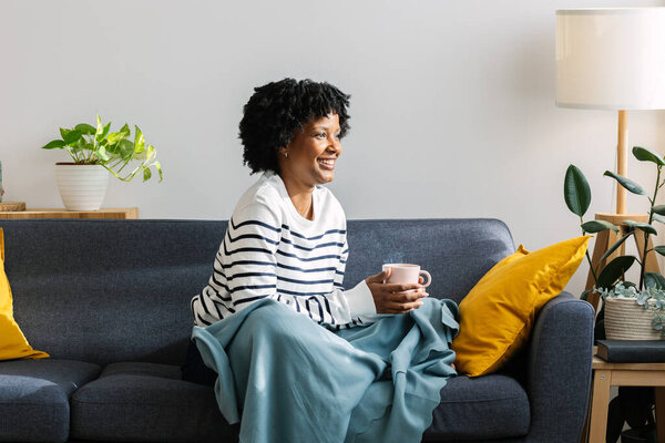 Молодая черная женщина наслаждается спокойным моментом на диване с теплым горячим чаем или кофейным напитком, завернутая в уютное одеяло в своей гостиной в зимний день