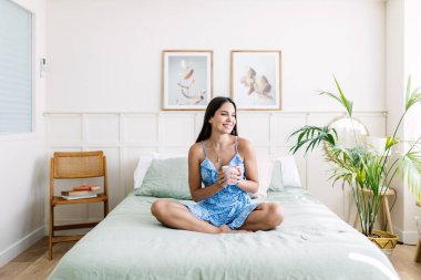 Serene young woman enjoying a quiet moment in her bedroom, savoring a warm beverage while sitting comfortably on her bed