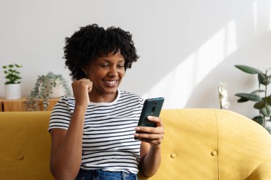Happy young black girl celebrating good news after read message on phone sitting on yellow sofa at modern apartment