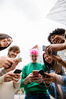 Vertical photo of diverse young friends having fun using mobile phones at city street. Millennial generation people laughing while watching social media content on cellphone app