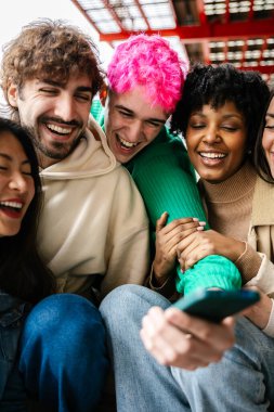 Vertical shot of young students bonding on campus steps, laughing while using phone to browse social media. Friendship and leisure concept.