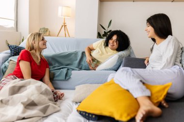 Three cheerful young women are enjoying a slumber party, chatting and laughing together on a comfortable sofa. Female friendship and youth concept