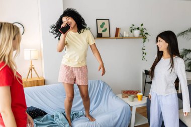 Three cheerful young women enjoying a slumber party, singing and dancing on a sofa in a cozy home environment. Female friendship and youth concept