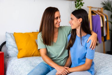 Mid adult mother and her 15-year-old teenage student daughter are sitting together on the bed, hugging warmly in a cozy bedroom. Family love, emotional support, and parenting care concept.