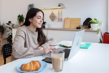 Dizüstü bilgisayarda çalışan genç üniversiteli kadın mutfaktaki masada oturuyor. E-öğrenim ve eğitim yaşam tarzı konsepti