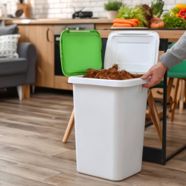 Bir birey, taze sebzelerle çevrili bir apartmanın içindeki gübre konteynırına organik atık ekler ve organizasyona duyulan ihtiyacı vurgulayan dağınık bir mutfak ortamı.