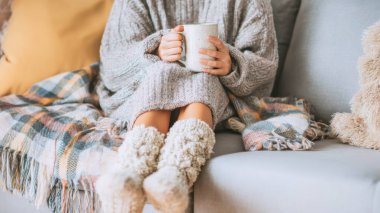 Genç bir kadın evdeki rahat bir kanepede oturuyor, yumuşak bir örgü kazağa sarılmış ve sıcak bir içecek tutuyor. Etrafı rahat battaniyelerle çevrili. Rahat ve davetkar bir atmosfer yaratıyor..