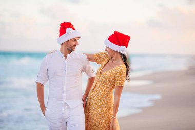 Young romantic couple in red Santa hats sitting on tropical white sand beach celebrating Christmas