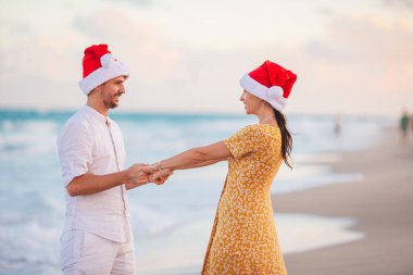 Young romantic couple in red Santa hats sitting on tropical white sand beach celebrating Christmas
