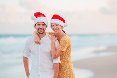 Young romantic couple in red Santa hats sitting on tropical white sand beach celebrating Christmas