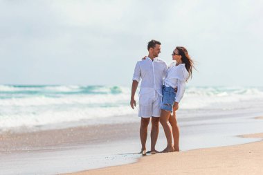 Kumsal tatilinde birbirine aşık mutlu çift gün batımında birlikte yürüyor. Beyaz elbiseli genç kadın ve beyaz şortlu beyaz adam..