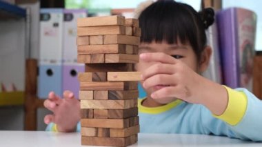 Cute little girl playing with wooden game (jenga) on a table in a room at home. Asian child playing wood blocks stack game indoors. Learning and development of fine motor skills.