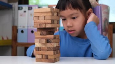 Cute little girl playing with wooden game (jenga) on a table in a room at home. Asian child playing wood blocks stack game indoors. Learning and development of fine motor skills.