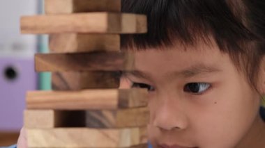 Cute little girl playing with wooden game (jenga) on a table in a room at home. Asian child playing wood blocks stack game indoors. Learning and development of fine motor skills.
