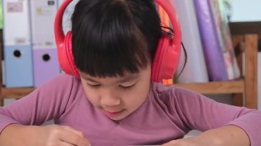 Cheerful little girl in headphones singing and drawing with colored pencils on paper sitting at table in her room at home. Creativity and development of fine motor skills.
