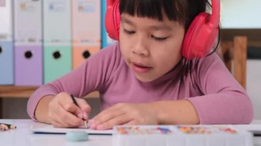 Cheerful little girl in headphones singing and drawing with colored pencils on paper sitting at table in her room at home. Creativity and development of fine motor skills.