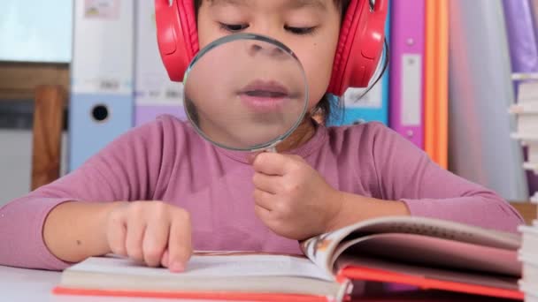 Весела Маленька Дівчинка Навушниках Читає Книгу Збільшувальним Склом Сидячи Столом — стокове відео