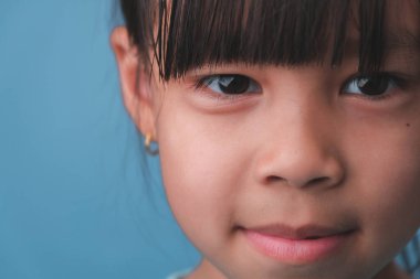 Kameraya bakan gülümseyen küçük bir kızın portresi. Mavi arka planda poz veren sevimli Asyalı kız yüzünü kapat..
