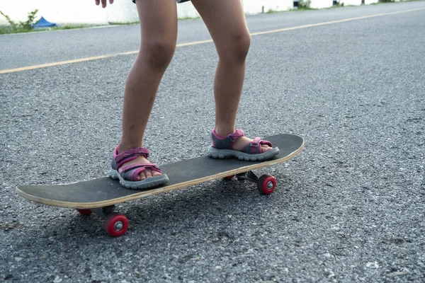 Cute little girl learns how to skateboard at outdoor street, focus on legs standing on board. Healthy sports and outdoor activities for school children in the summer.