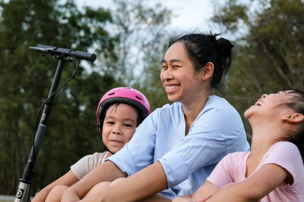 stock image Mother and children sit and rest after riding scooters in the park. Mother and daughters spend their free time riding scooters outdoors together. Happy Loving Family.