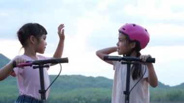 Yaz bahçesinde gülümseyen ve poz veren iki küçük kız. Parkta scooter süren mutlu çocuk. Çocuklar için sağlıklı spor ve açık hava aktiviteleri.