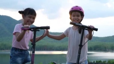 Yaz bahçesinde gülümseyen ve poz veren iki küçük kız. Parkta scooter süren mutlu çocuk. Çocuklar için sağlıklı spor ve açık hava aktiviteleri.