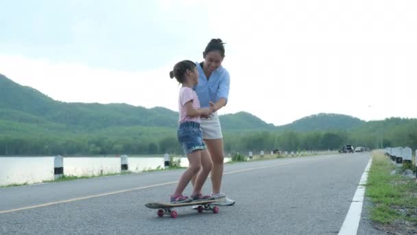 母は彼女の娘に公園でスケートボードの方法を教える 子供のスケートボードに乗る 夏の学校の子供たちのための健康的なスポーツや屋外活動 — ストック動画