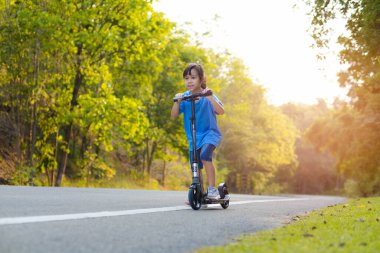 Aktif küçük kız bir yaz günü parkta scooter 'a biniyor. Kask takan sevimli küçük kız açık hava parkında scooter sürmekten hoşlanıyor. Çocuklar için boş zaman aktiviteleri ve açık hava sporları.