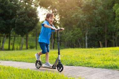 Aktif küçük kız bir yaz günü parkta scooter 'a biniyor. Kask takan sevimli küçük kız açık hava parkında scooter sürmekten hoşlanıyor. Çocuklar için boş zaman aktiviteleri ve açık hava sporları.