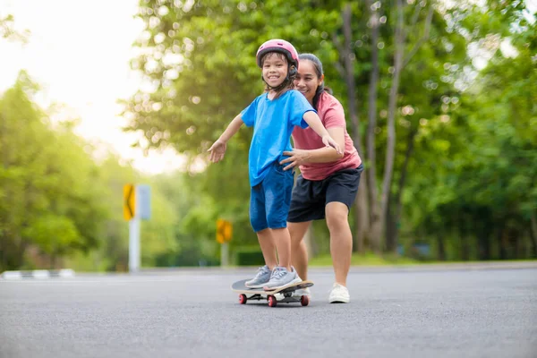 stock image Active little girl and mom enjoy skateboarding. Cute little girl wearing helmet practicing skateboarding in park. Mother trains her daughter to skateboard. Outdoor sports for children.