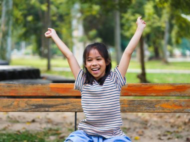 Parktaki bankta oturan mutlu küçük bir kız. Kollarını açmış gülümseyen küçük bir kız. Temiz hava soluyor. Sabahları doğanın tadını çıkarıyor..