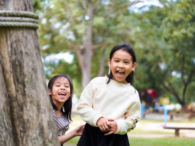 Ağacın arkasına bakan şirin küçük kız kardeşler güneşli bir bahçede dikiz oyunu oynayarak eğleniyorlar. Parkta gülümseyen çocuk