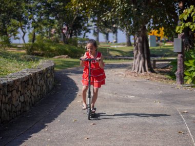 Güneşli bir yaz gününde parkta scooter süren tatlı bir kız. Çocuk için aktif eğlence ve açık hava sporu.