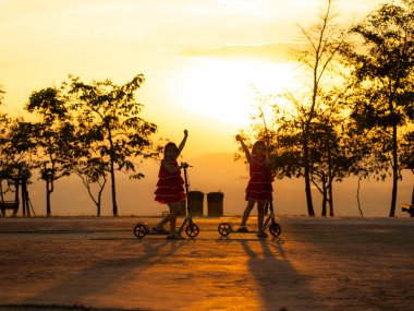 Parkta gün batımında parkta scooter süren şirin kız kardeşlerin silueti. Çocuk için aktif eğlence ve açık hava sporu.