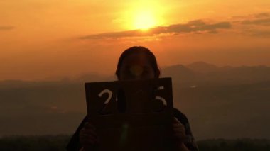 Günbatımı arka planında 2025 tabelasını tutan kadın silueti. Mutlu yıllar 2025.