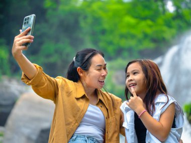 Bir dişi turist ve küçük tatlı kızı akıllı telefonlarıyla çarpıcı bir şelalenin yanında selfie çekiyorlar..