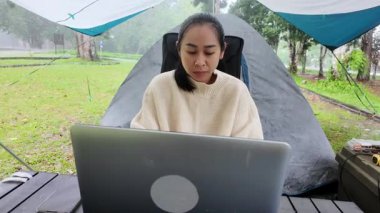 Asyalı bir kadın yağmur mevsiminde ulusal parkta kamp yaparken bilgisayarını uzaktan çalışmak için kullanıyor. Çalışma ve rahatlama konsepti.