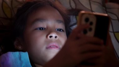 Asyalı kız cep telefonuna düşük ışıkla bakıyor ve telefonun mavi ışığı gözlerine zararlı. Tatlı kız gece geç saatte kanepede akıllı telefona bakıyor, çocuklarda ekran bağımlılığı..