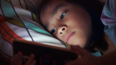 Asyalı kız cep telefonuna düşük ışıkla bakıyor ve telefonun mavi ışığı gözlerine zararlı. Tatlı kız gece geç saatte kanepede akıllı telefona bakıyor, çocuklarda ekran bağımlılığı..