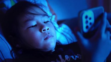 Asyalı çocuk geceleri akıllı telefon kullanarak yatakta yatıyor, ekran ışığı aydınlatma yüzü, ekran zamanı konsepti ve çocuklarda dijital bağımlılık
