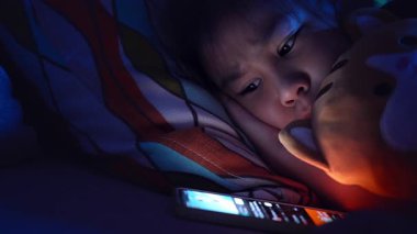 Asyalı çocuk geceleri akıllı telefon kullanarak yatakta yatıyor, ekran ışığı aydınlatma yüzü, ekran zamanı konsepti ve çocuklarda dijital bağımlılık