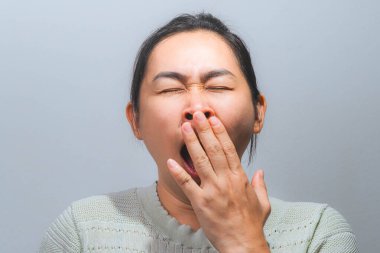 Asyalı bir kadın eliyle ağzını kapatıp esniyor. Sade bir arka planda yorgunluk ve yorgunluk gösteriyor..