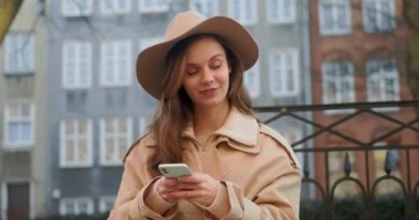 Şehir binalarında telefon kullanırken yürürken sevimli çekici bir kadın etrafına bakıyor güneş ışığında mutlu bir gülücük atıyor. Portre, genç, internet, cep telefonu, sokak, güzel, seyahat, mobil, şık. Yavaş