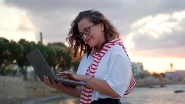 Young european women by the sea, working with a laptop on a sunset background.