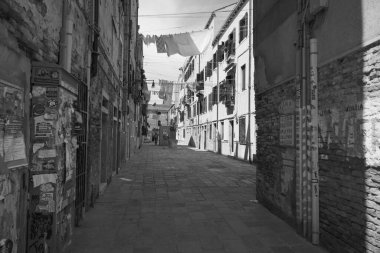 Venedik 'te dar bir sokak aralığının siyah-beyaz görüntüsü, tarihi binaların sergilenmesi, çamaşırların asılması, ve Venedik' te parlak bir gökyüzünün altında dokulu taş bir cadde.