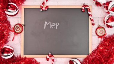 Beyaz arka planda kırmızı süslemeler olan karatahtaya Mutlu Noeller yazıyorum. Noel tebrik kartı düzdü. Hareketi durdur
