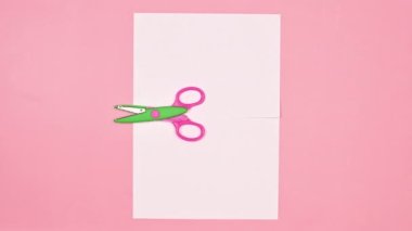 Renkli makas pastel pembe arka planda beyaz kağıt kesiyor. Hareketi durdur. Düz yatıyordu